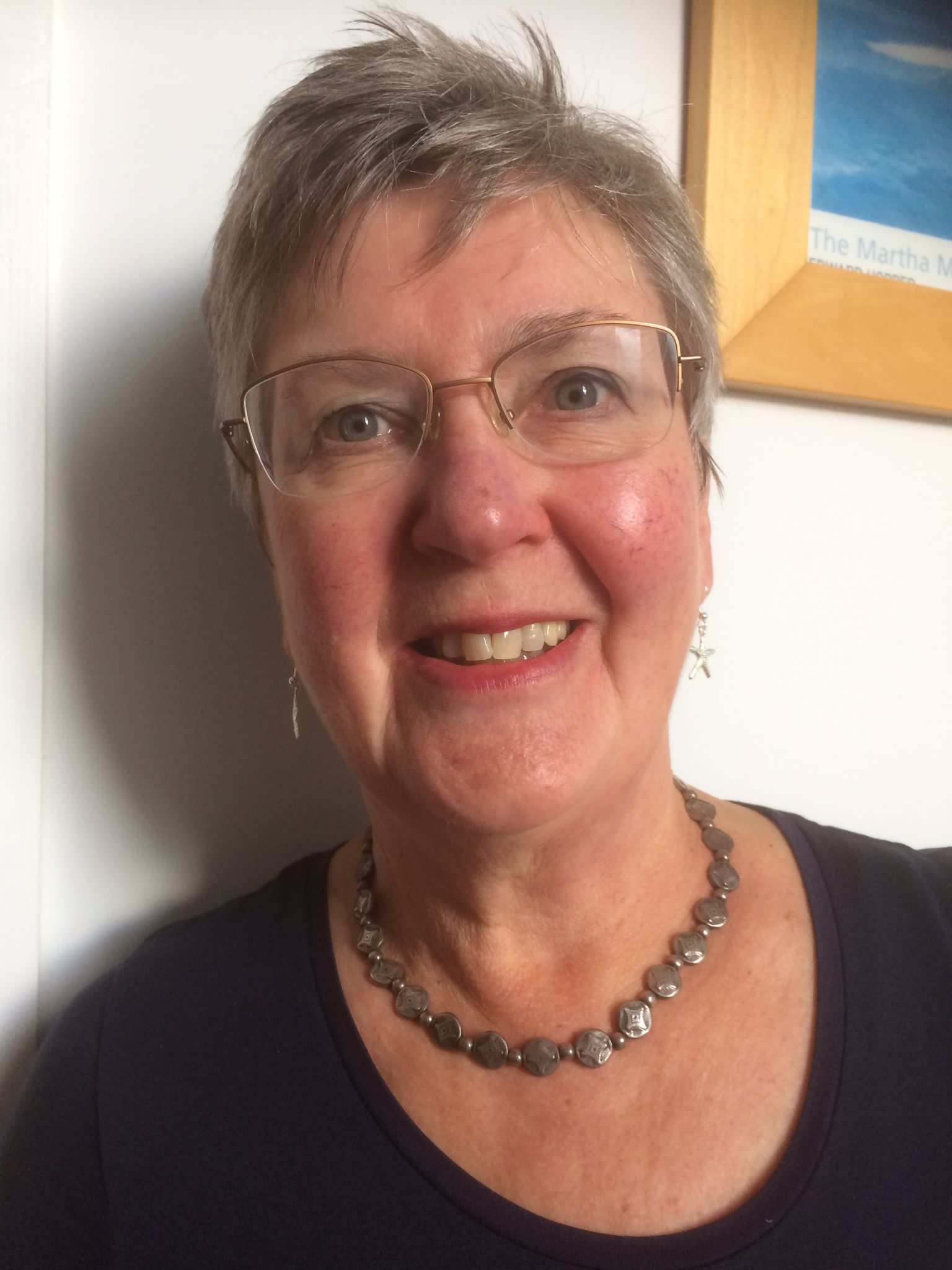 Picture shows a portrait photograph of Jill Carr, author of Astrolations! Jill is a white woman, in her 70s. She has short, ash blonde hair and is smiling looking at the camera. Jill is wearing glasses, dangly earrings and a chunky silver necklace. 